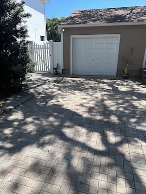 A residential driveway leading to a garage with a white door, surrounded by a white picket fence, potted plants, and a brick pathway.