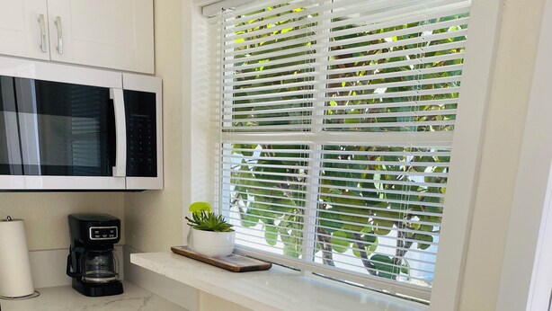 A kitchen with white cabinets, a microwave, a coffee maker, two wooden stools, and a window with blinds.