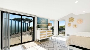 Une chambre à coucher moderne avec une grande porte-fenêtre coulissante donnant sur une terrasse, une commode blanche, un lit avec une tête de lit blanche et une vue sur l’extérieur.