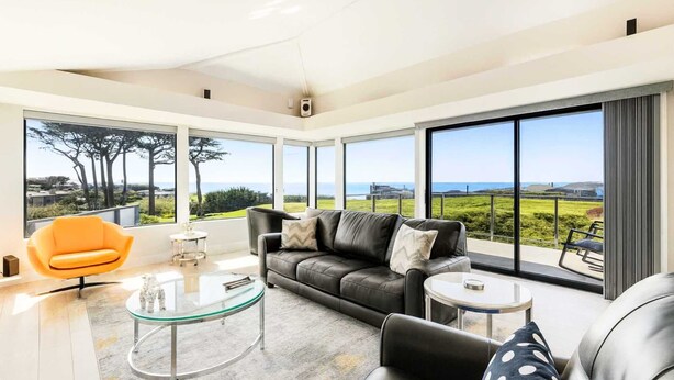 Un salon moderne avec un canapé en cuir, une table basse en verre et un fauteuil orange vif. De grandes fenêtres offrent une vue sur la campagne et la mer.