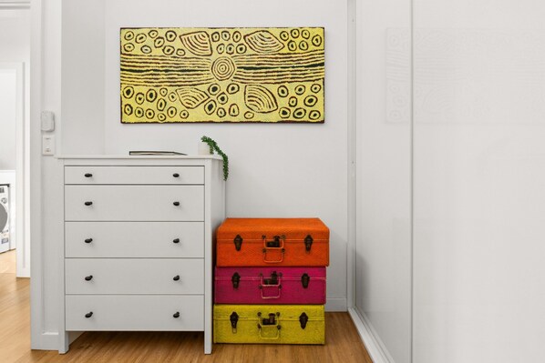 A white dresser with drawers, a wall-mounted artwork, and three stacked suitcases in orange, pink, and yellow. - Luxury 2-bed With Ocean Views and Garden (Sydney)