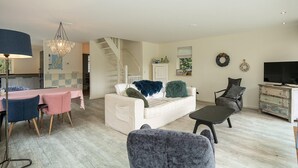 Een woonkamer met een witte bank, een grijze fauteuil, een eettafel met roze en blauwe stoelen, een kroonluchter en een trap naar een andere kamer.