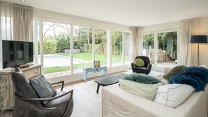Een ruime woonkamer met een grote televisie, comfortabele zitmogelijkheden en uitzicht op de tuin door grote ramen.
