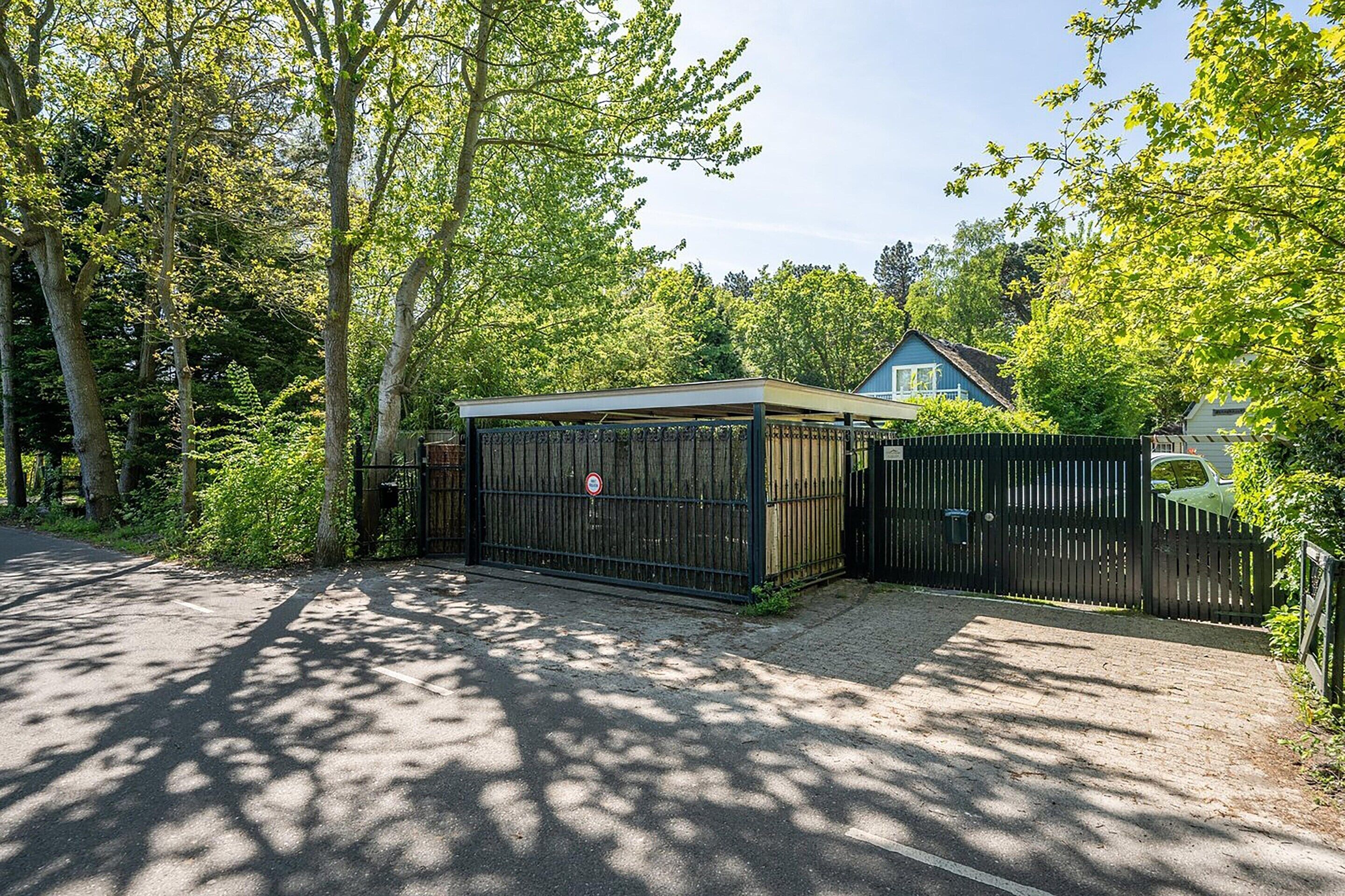 A gated parking area with a covered shelter, surrounded by trees and a house in the background.