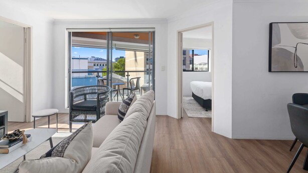 Una sala de estar moderna con sofá, una mesa de centro y televisor. Hay un balcón con vista a edificios y una piscina.