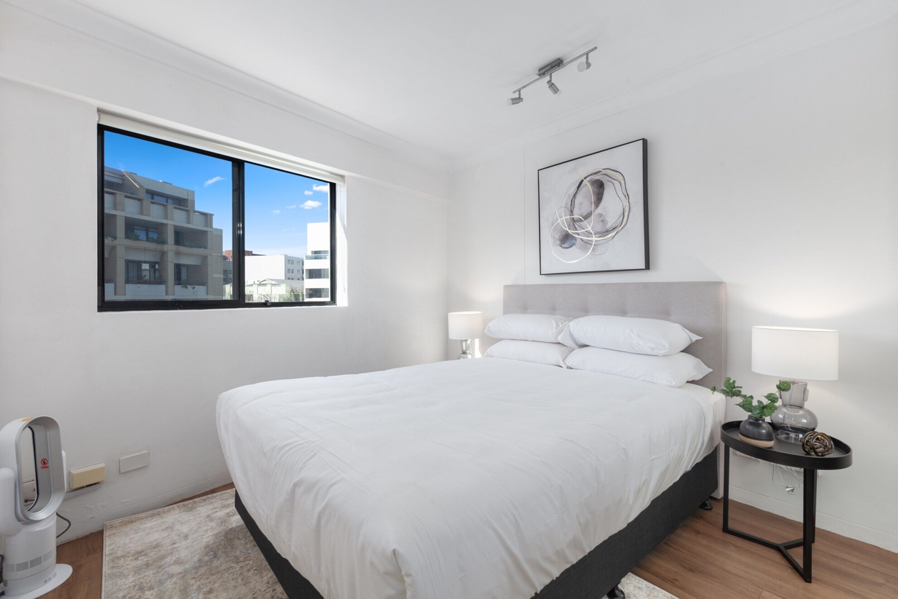 Un dormitorio moderno con una cama grande, una mesita de noche con lámparas, una obra de arte en la pared y una ventana con vistas a edificios.
