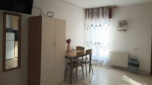 Une petite pièce bien éclairée, avec une table à manger, des chaises, un téléviseur fixé au mur, un radiateur et une poubelle.