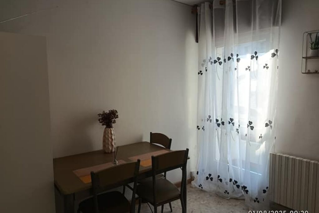 Ein Essbereich mit Tisch, Stühlen, einer Vase mit Blumen, transparenten Vorhängen mit Blumenmuster und einem Heizkörper an der Wand.