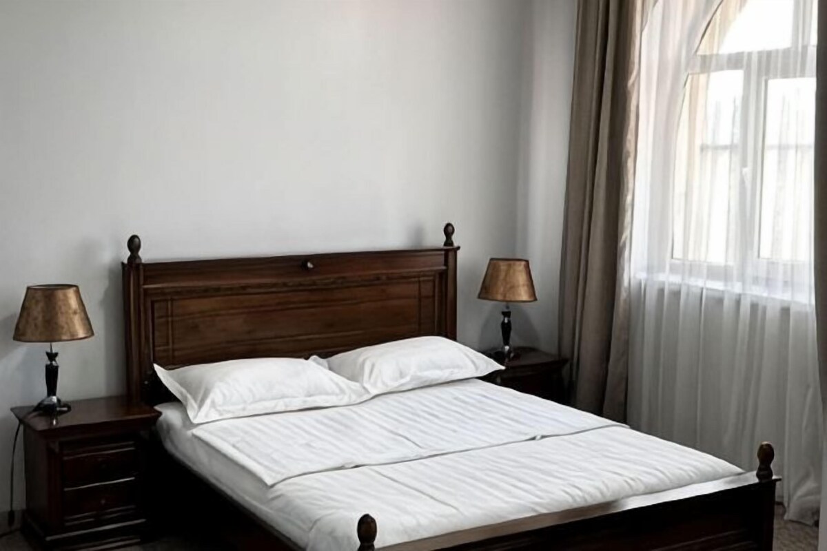 A neatly made bed with white linens, two bedside lamps, and a wooden headboard in a room with a plain wall and a window with curtains.