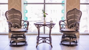 Two wicker chairs with a small table and a vase of flowers between them, positioned in front of large windows with a view of buildings. - Shah Palace Hotel (Bishkek)
