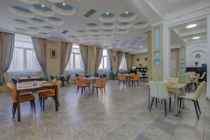Ein geräumiger Essbereich mit mehreren Tischen und Stühlen, einer gemusterten Decke und großen Fenstern mit Vorhängen.