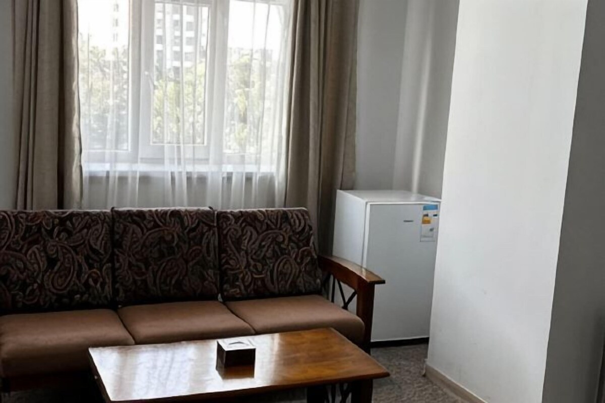 A room with a sofa, a small table, a refrigerator, and a window with curtains.