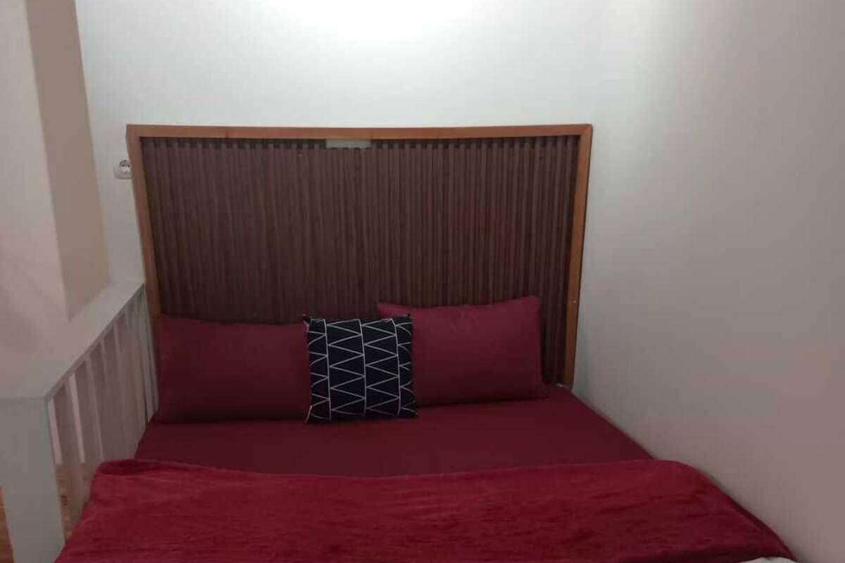 Una cama individual con cabecera de madera, ropa de cama roja y blanca, y una almohada estampada.