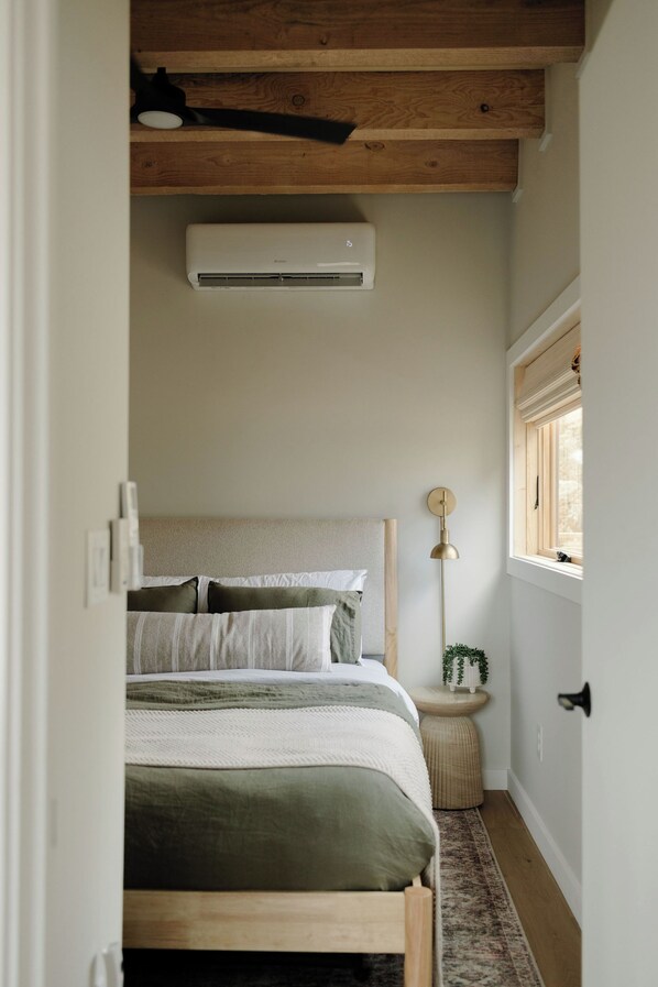 Un dormitorio con una cama de madera, una lámpara de noche, una mesita redonda con una planta y un equipo de aire acondicionado en la pared.