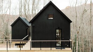 Une cabane en bois noire avec un balcon, une petite dépendance attenante et une vue sur une zone boisée.