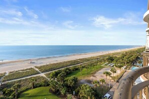 A beachfront view from a balcony, including a sandy beach, ocean, and a well-maintained lawn with palm trees. - Oceanfront King, Walk-in-Shower, Fireplace, Heated Pools, Exc Walkability (Myrtle Beach)