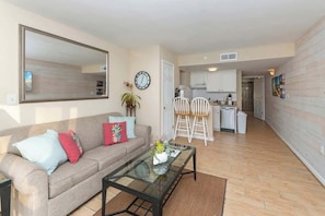 A modern living room with a sofa, coffee table, and a large mirror. A kitchen is visible in the background with white cabinets and a dining area. - Oceanfront King, Walk-in-Shower, Fireplace, Heated Pools, Exc Walkability (Myrtle Beach)