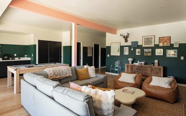 A modern living room with a sofa, coffee table, and bean bag chairs. There is a pool table and a kitchen area in the background.