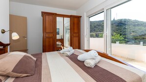 A hotel room with a large bed, a wooden wardrobe, a bathroom with a mirror, and a balcony with a view of the hills. - Apartment Zelenika Villa by Villas Guide (Ploce)