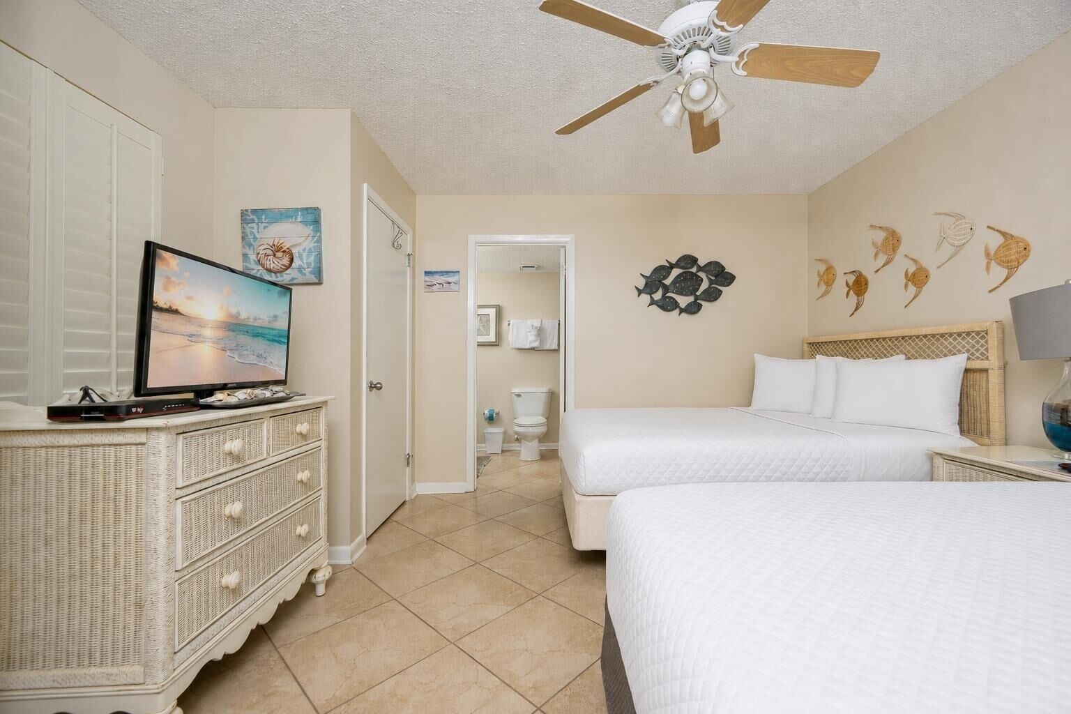 Habitación de hotel con televisión, dos camas, ventilador de techo y baño visible a través de una puerta abierta.