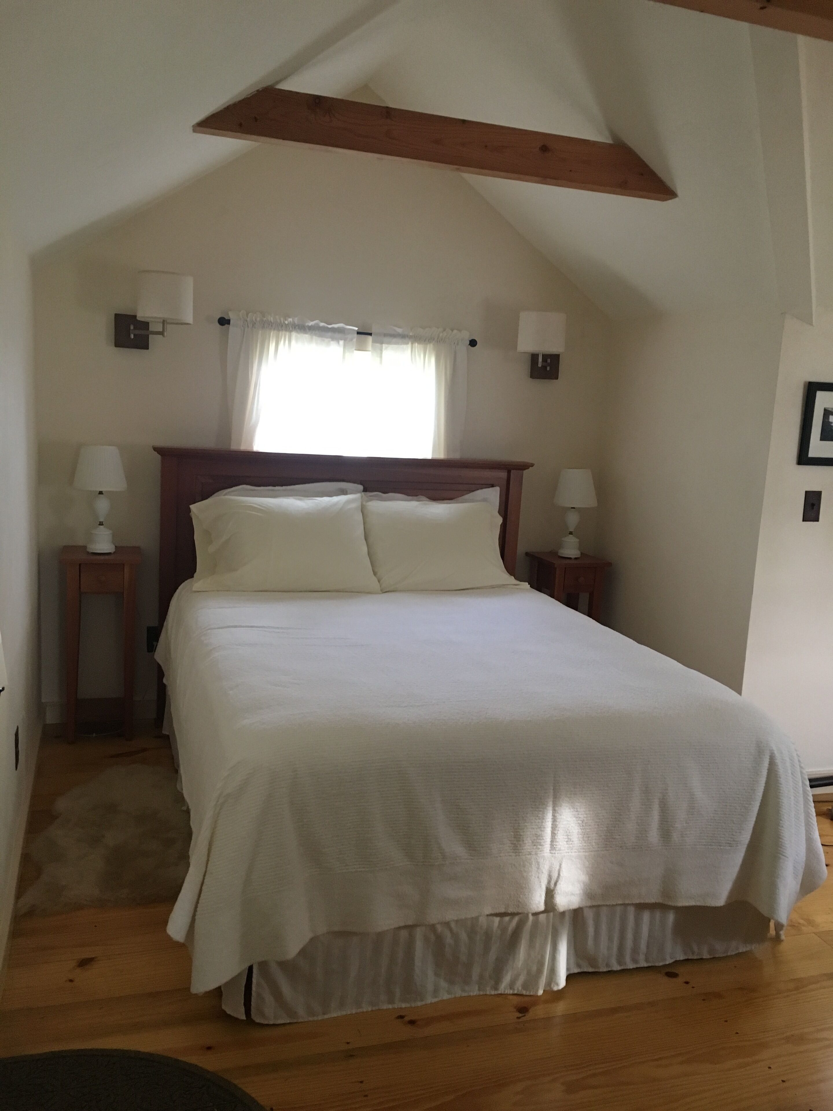 Un dormitorio con techo de madera, una cama grande con ropa de cama blanca, dos mesitas de noche con lámparas y una ventana con cortinas.
