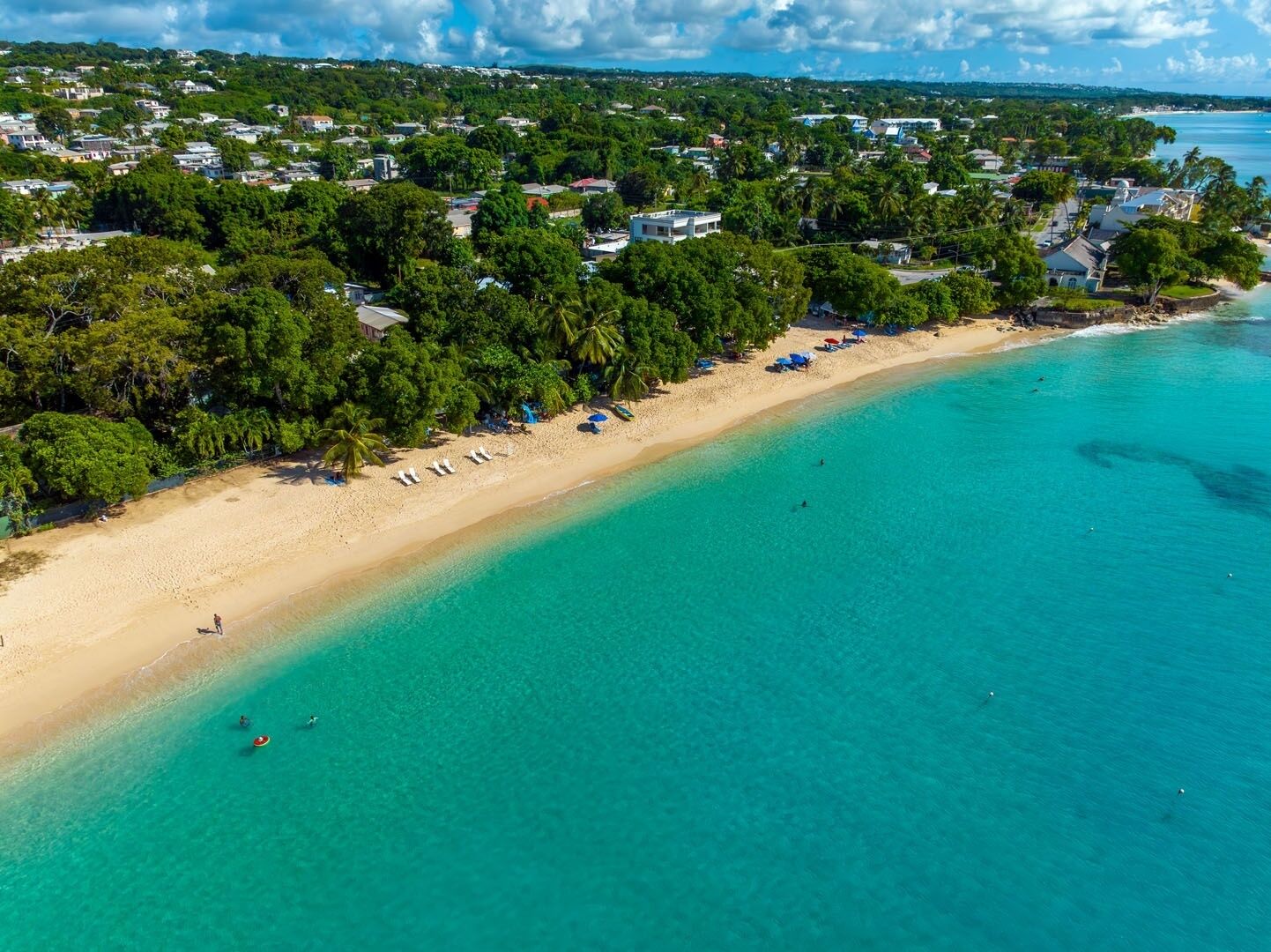 An aerial view of a beach with clear turquoise waters, a sandy shore, and a backdrop of residential buildings.