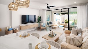 A modern living room with a flat-screen TV, wicker pendant lights, a dining area with a table set for two, and a sliding glass door leading to an outdoor seating area.
