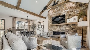 A modern living room with a stone fireplace, a dining area, and a large window offering a view of trees. - 37 Alcove 3 Bedroom Townhouse (Keystone)
