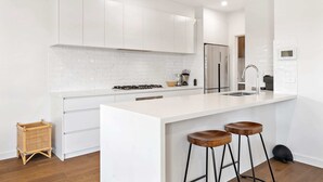 A modern kitchen with white cabinetry, a large island with bar stools, and a built-in oven. - Bright 2-bed Glenroy Home With Courtyard (Melbourne)