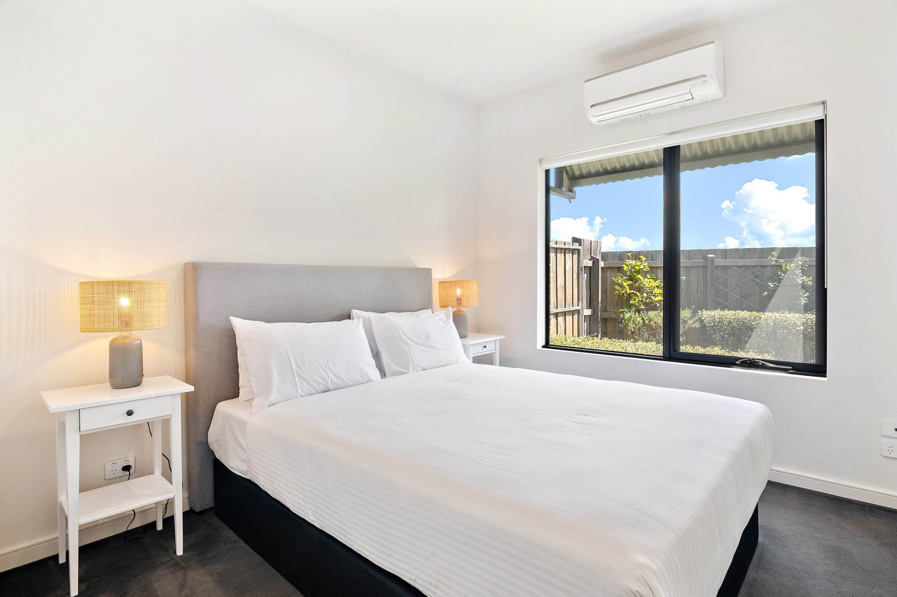A bedroom with a large bed, white nightstands, a lamp, and a window with a view of the outside.
