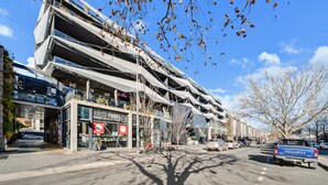 Een modern meerlaags gebouw met een glazen gevel, een Naked Foods winkel en een geparkeerde Isuzu vrachtwagen.