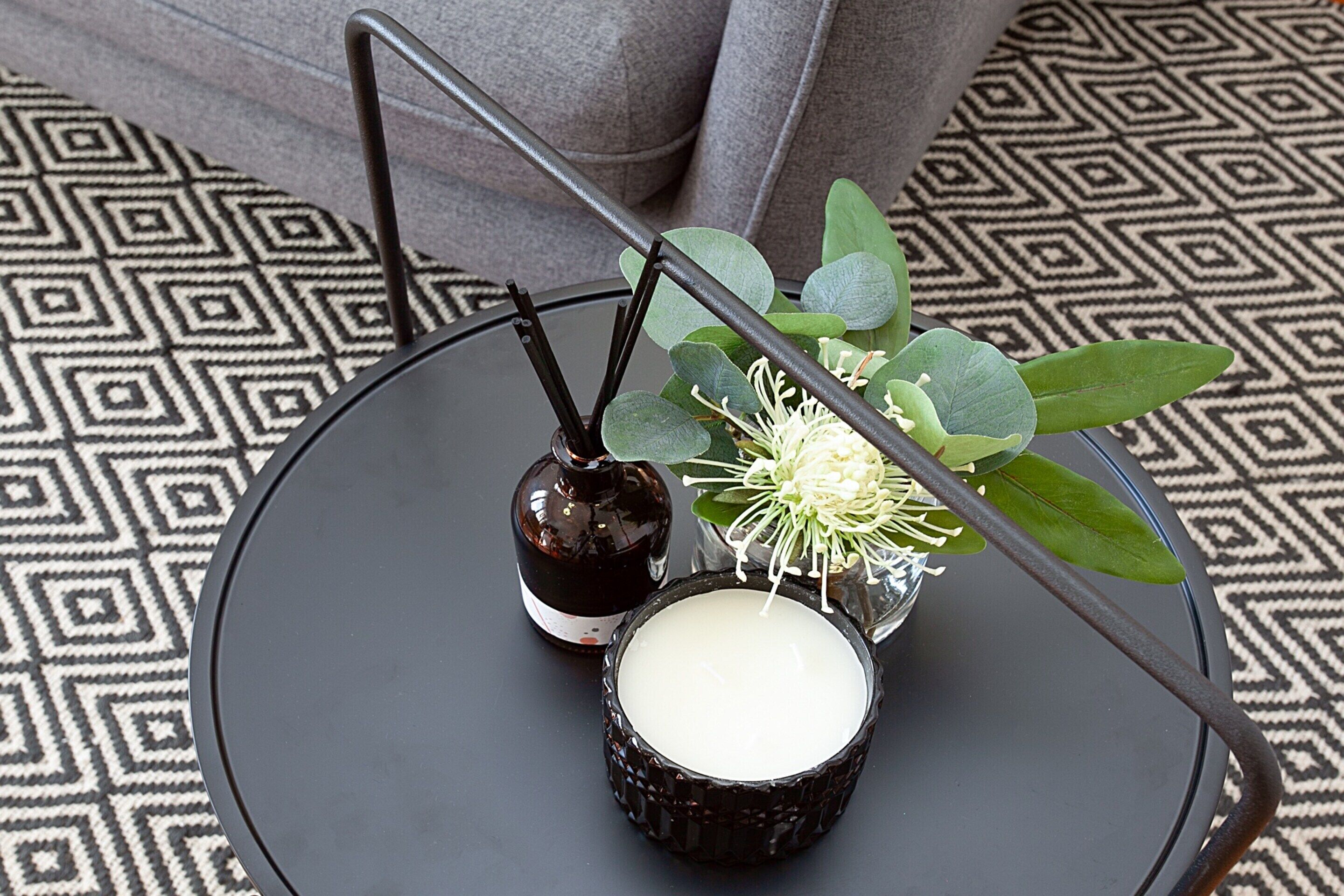 Un plateau noir sur lequel se trouvent une bougie, une bouteille et un petit vase contenant des feuilles vertes et des fleurs blanches, le tout posé sur un sol à motifs.
