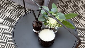 A black tray with a candle, a bottle, and a small vase with green leaves and white flowers on a patterned floor. - Perfect Located Comfy Studio With Balcony (Sydney)