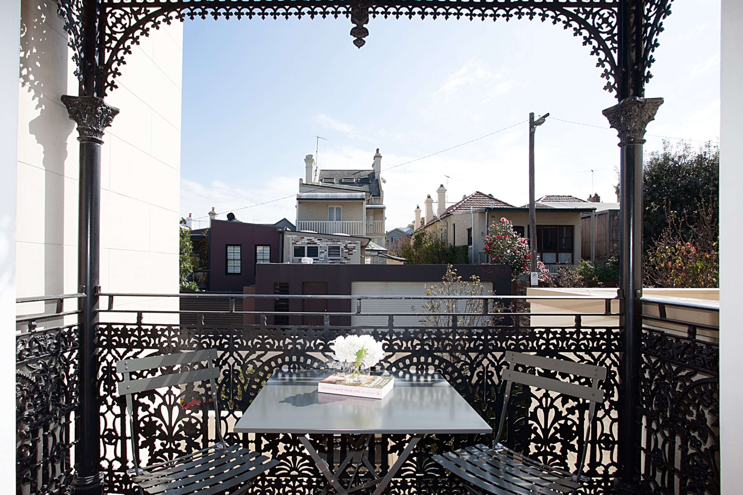 A balcony with a table and chairs, overlooking a street with buildings and a clear sky.