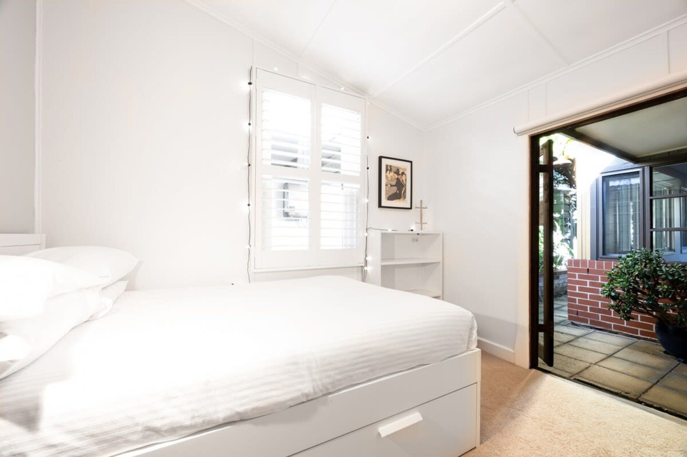 Een slaapkamer met een bed, een raam met zonwering, een klein nachtkastje met een fotolijstje en uitzicht buiten door een glazen deur.