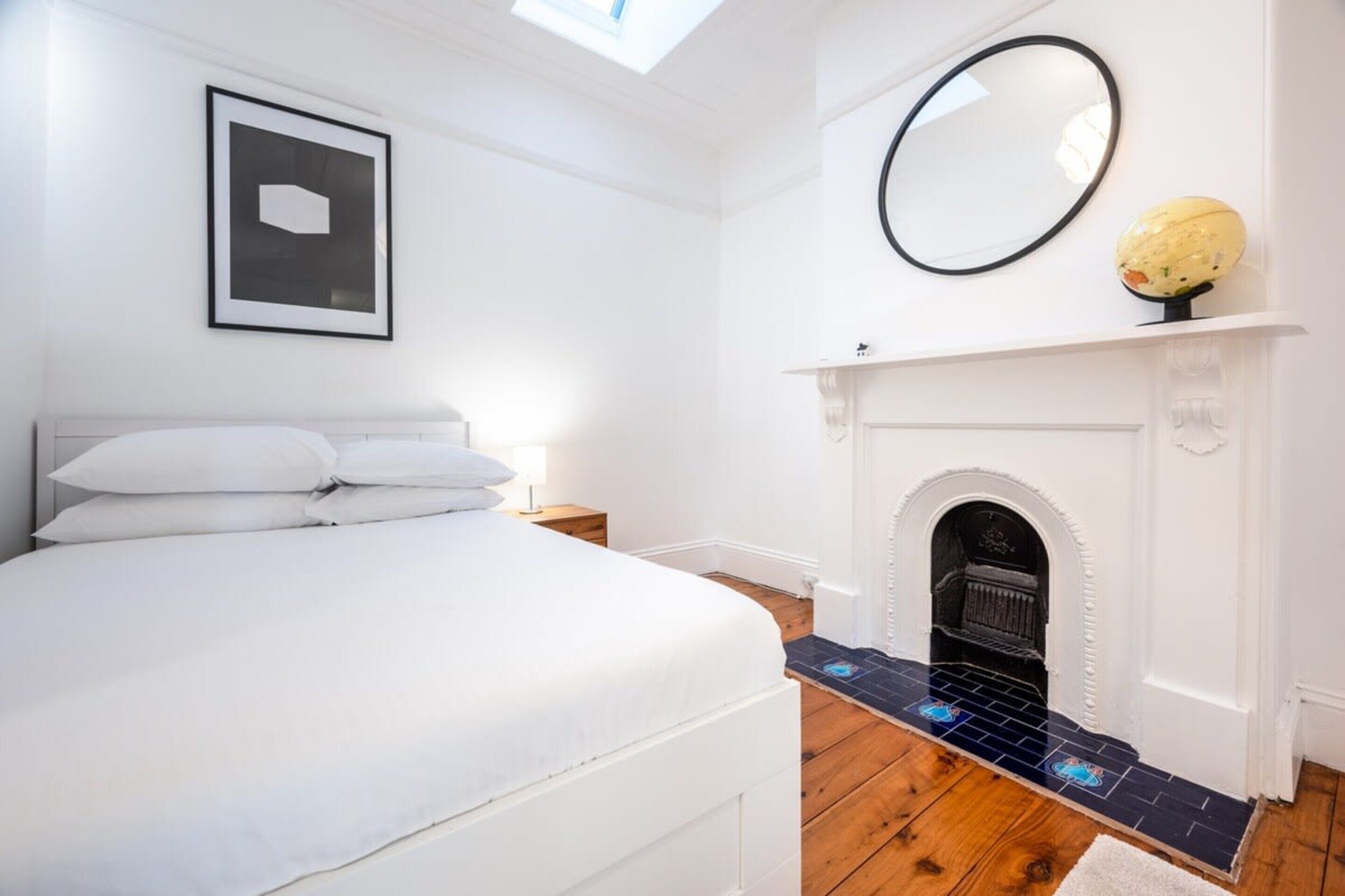 A bedroom with a bed, a fireplace, a framed picture, and a round mirror.