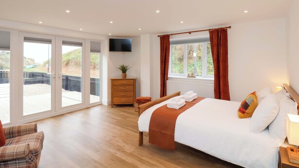 A bedroom with a bed, a TV, a window with curtains, a wooden dresser, and a sliding glass door leading to an outdoor area. - Sandy Nook (Redcar)