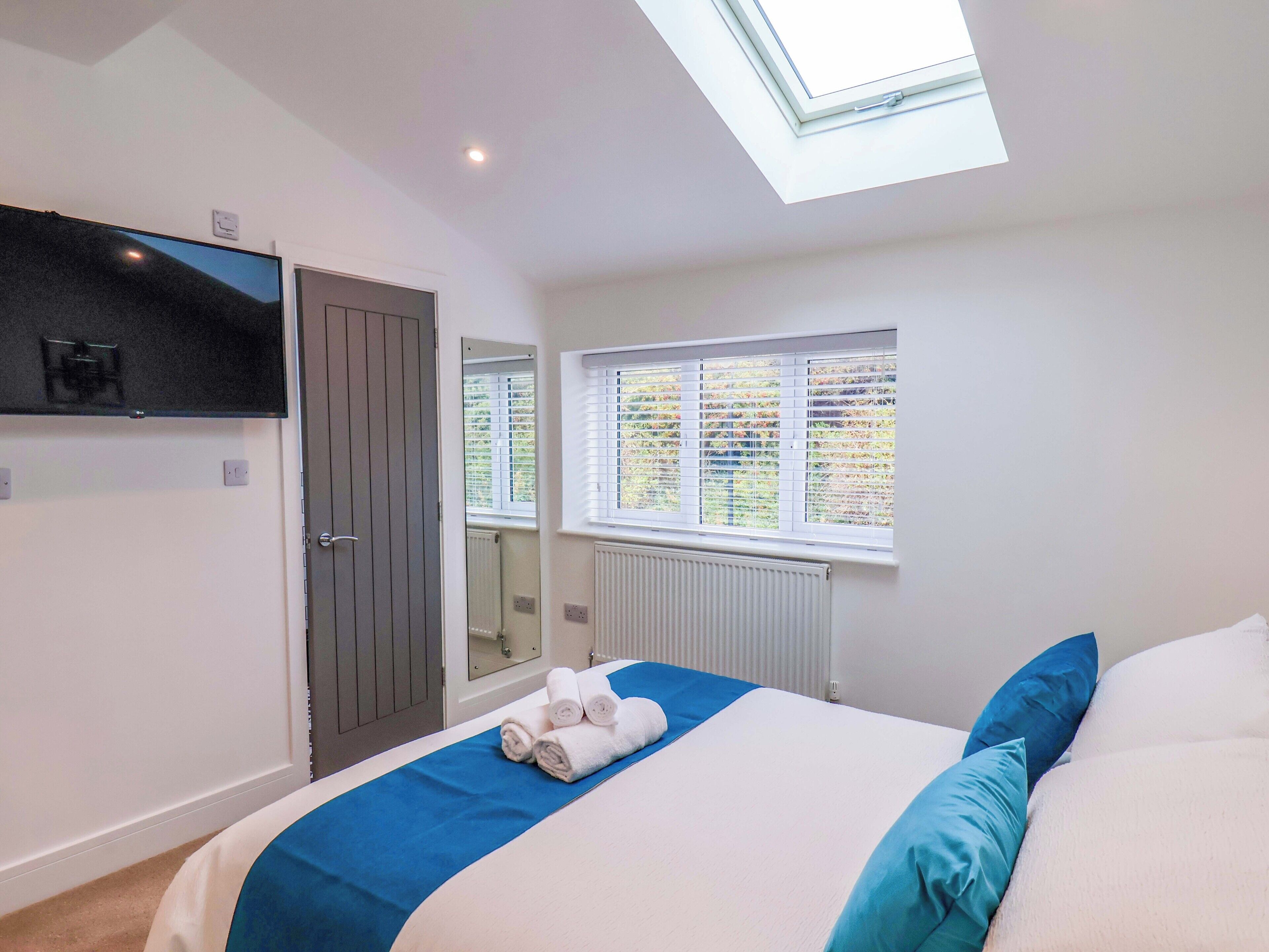 A bedroom with a skylight, a bed with blue and white bedding, a TV on the wall, and a window with blinds.