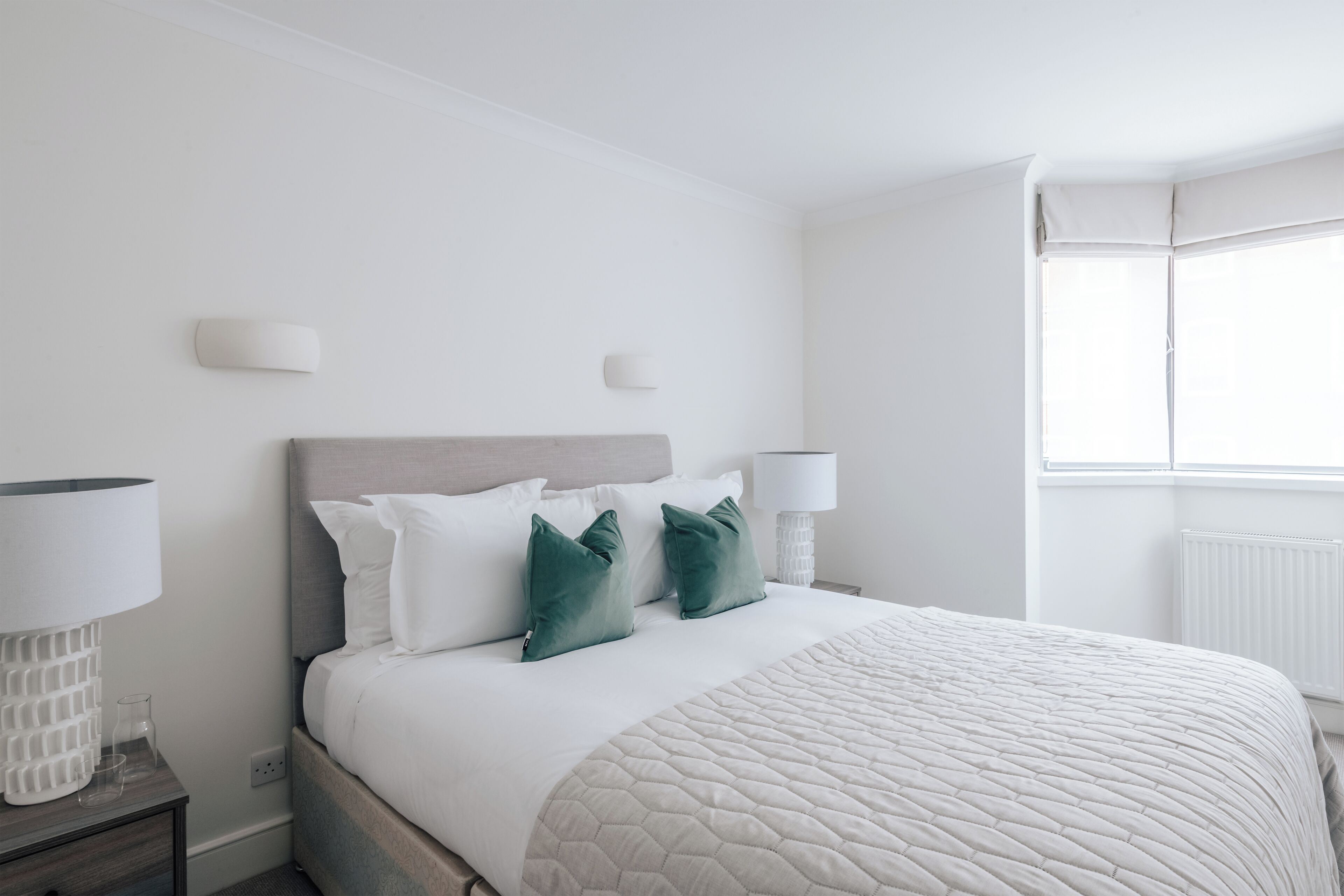 A neatly made bed with a quilted blanket, two green pillows, and a wooden headboard. There are two bedside lamps on a nightstand.