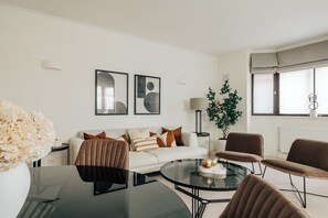 Una sala de estar moderna con un sofá blanco, cojines marrones y a rayas, una mesa de centro de vidrio y sillas marrones. En la pared hay dos cuadros abstractos enmarcados, una planta en maceta y una lámpara.