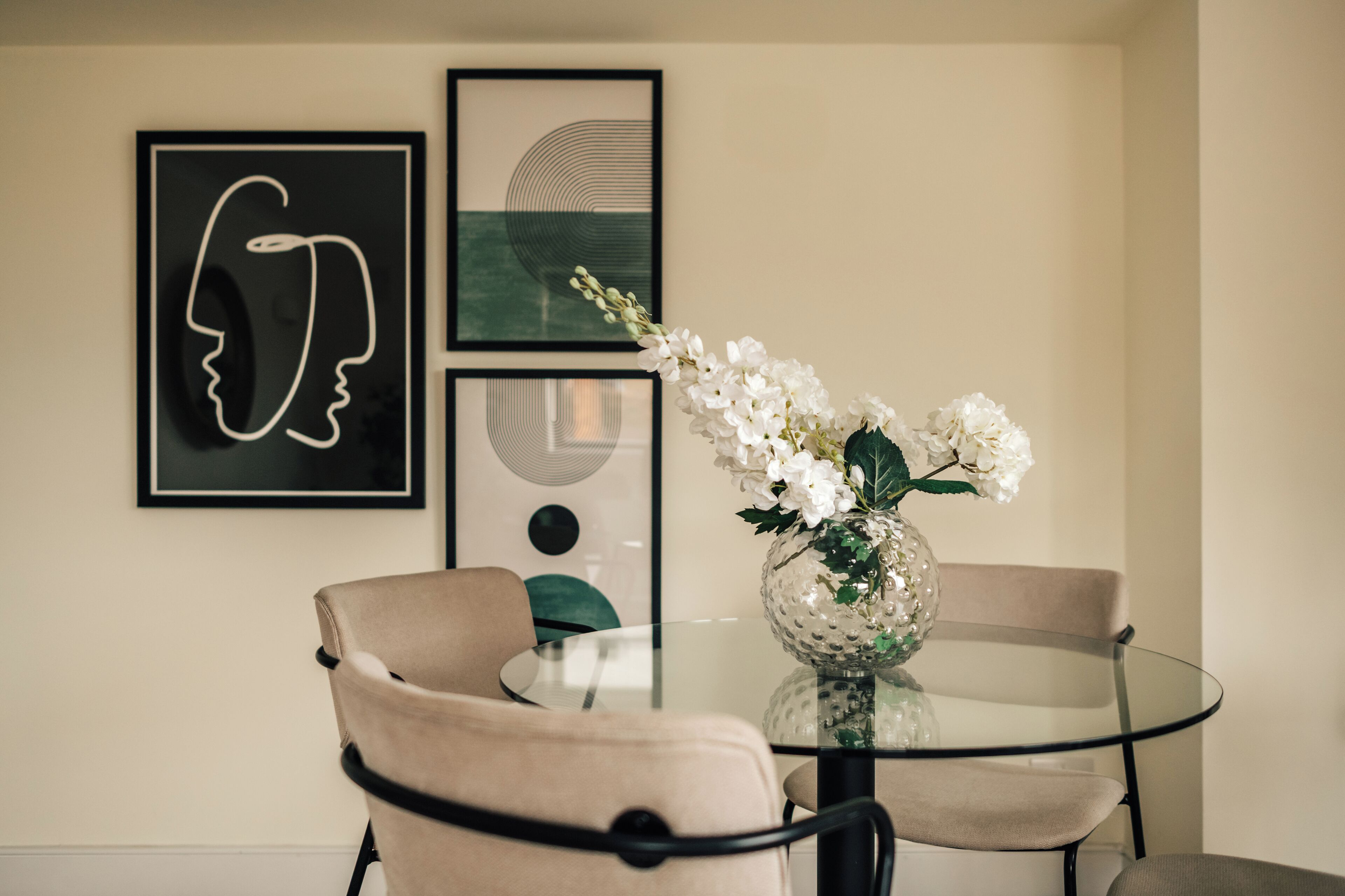 A dining area with a glass table, two beige chairs, and a vase with white flowers.
