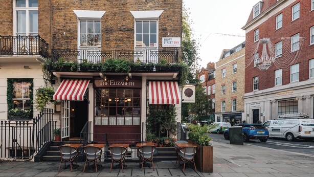 A brick building with a ground-floor shop named 'The Elizabeth' featuring a red and white striped awning, outdoor seating with wicker chairs, and a street sign indicating 'Ebury Street SW1'.