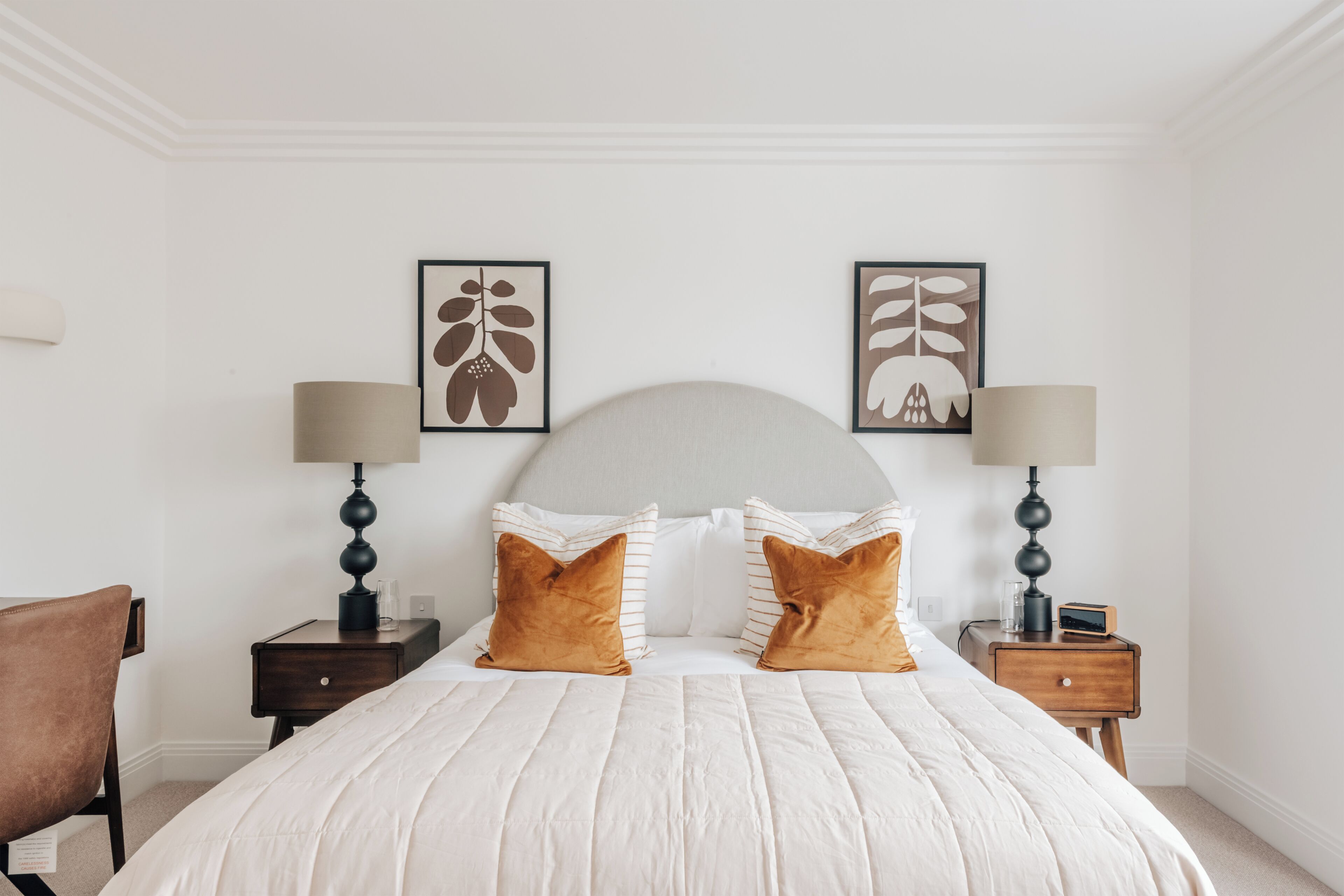 Un lit bien fait, avec une literie blanche et deux oreillers marron, flanqué de deux tables de chevet avec des lampes, et deux œuvres d’art encadrées accrochées au mur au-dessus du lit.