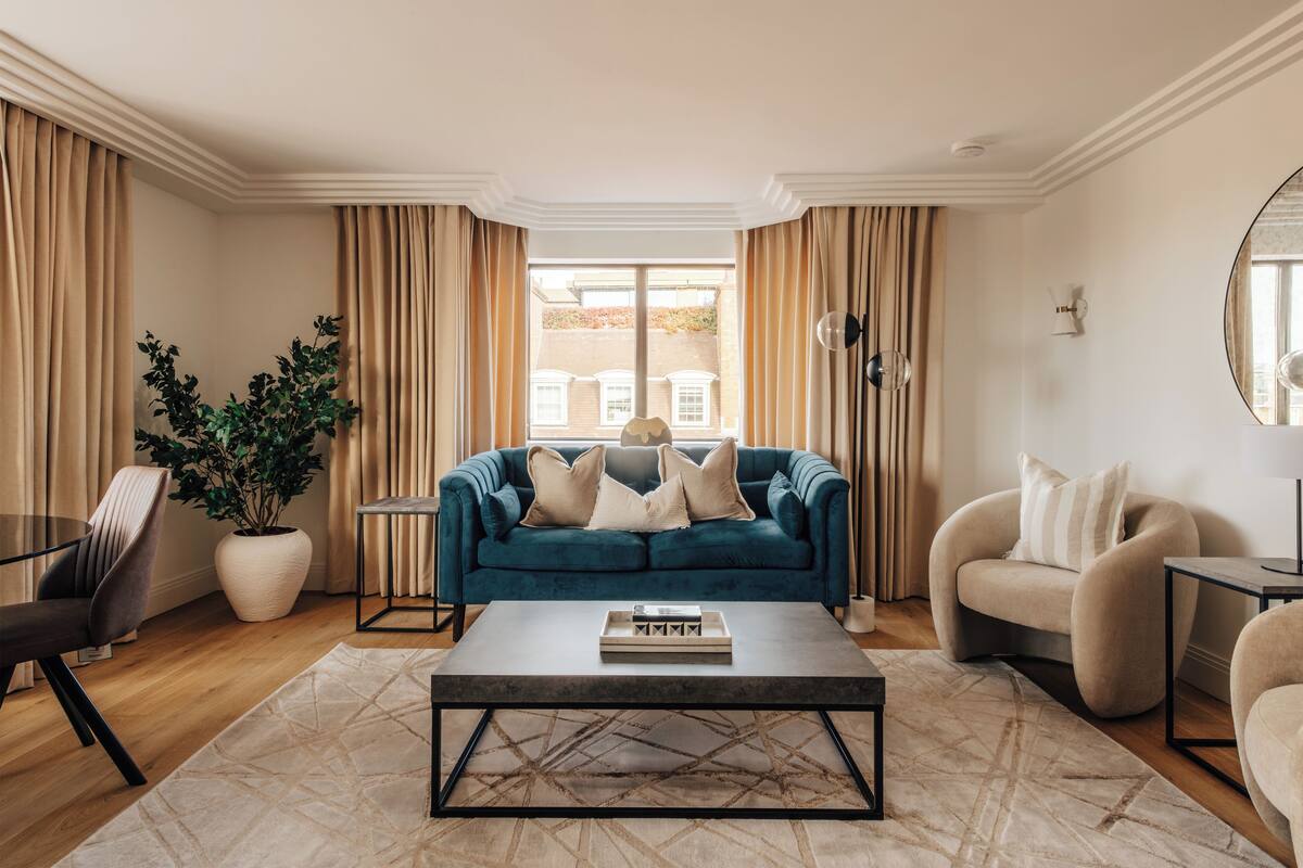 Un salon avec un canapé bleu, des fauteuils beige, une table basse et une grande fenêtre avec des rideaux.