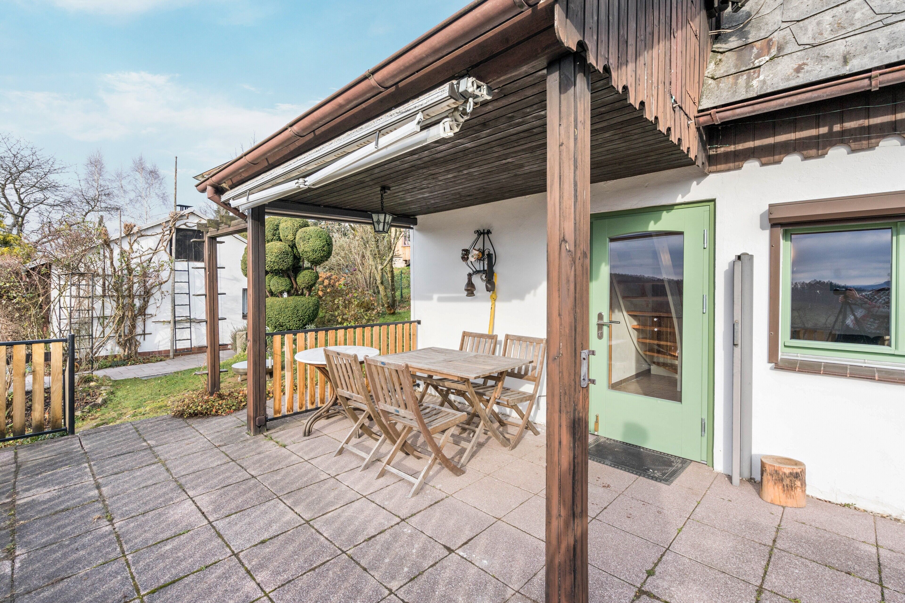 A patio with a wooden table and chairs, a green door, and a white wall with a window.