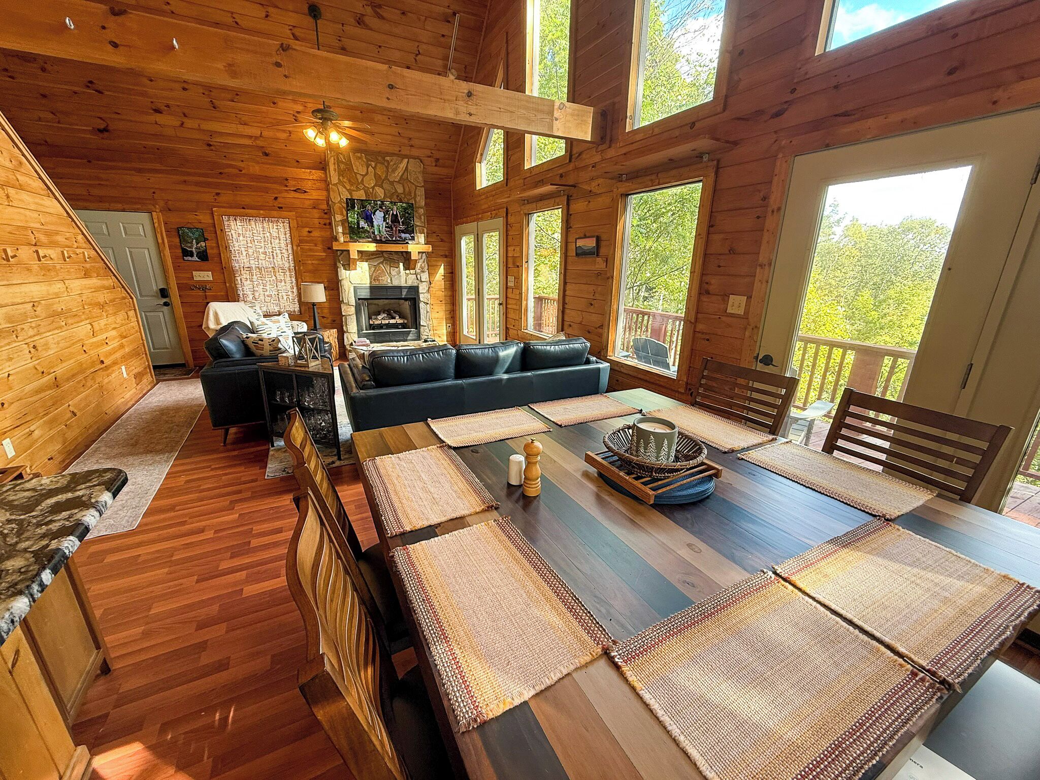 A cozy cabin interior with a stone fireplace, wooden furniture, and large windows offering views of the outdoors.