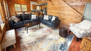 A cozy cabin interior with a living room featuring two black leather sofas, a coffee table, and a patterned rug. A staircase leads to an upper level, and a kitchen is visible in the background. - Mountaintop Memories-Hot Tub, Game Room, Pet Friendly, Near Grayson Highlands (Grassy Creek)