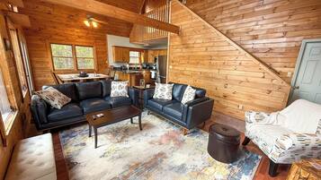 Een gezellig cabin interieur met een woonkamer, bestaande uit twee zwarte leren banken, een salontafel en een kamerkleed. Een trap leidt naar een hoger niveau, en er is een keuken in de achtergrond zichtbaar.