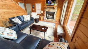 A cozy log cabin interior with a fireplace, a television mounted above it, a leather sofa, and a stone fireplace surround. - Mountaintop Memories-Hot Tub, Game Room, Pet Friendly, Near Grayson Highlands (Grassy Creek)