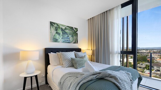 A modern hotel room with a large bed, a nightstand, a lamp, and a view of the city through large windows. - Surfers Paradise 2-bed Flat With Luxury Amenities (Gold Coast)
